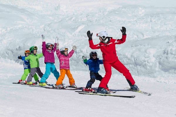 Young children in colorful gear learn to ski with an instructor on a sunny, snowy slope.