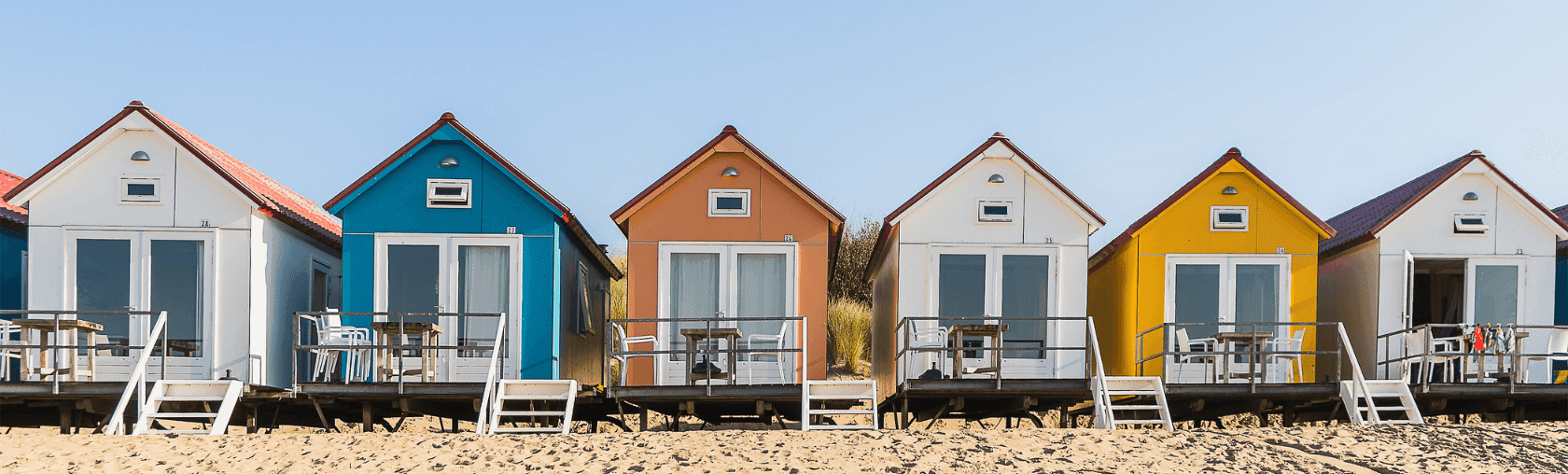 Row of vibrant beach huts with porches on a sandy beach against a clear sky.
